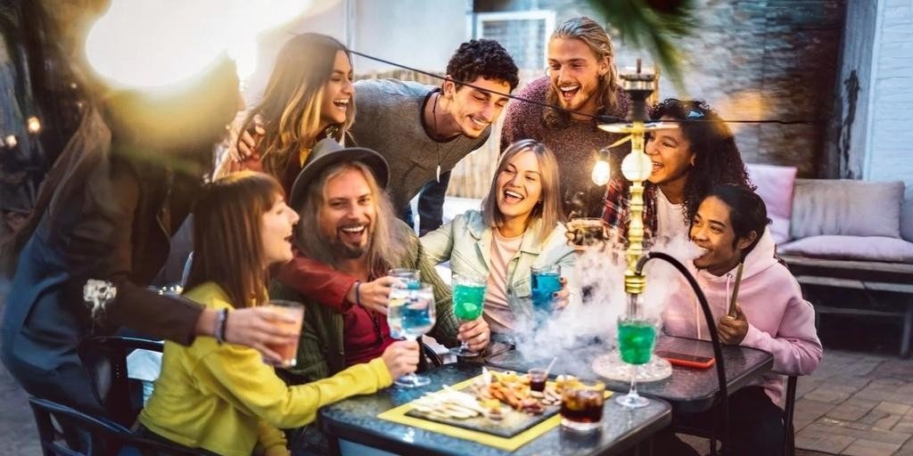 Group of friends laughing and sharing drinks around a table while enjoying shisha, with colorful beverages, food, and smoke creating a lively social atmosphere.