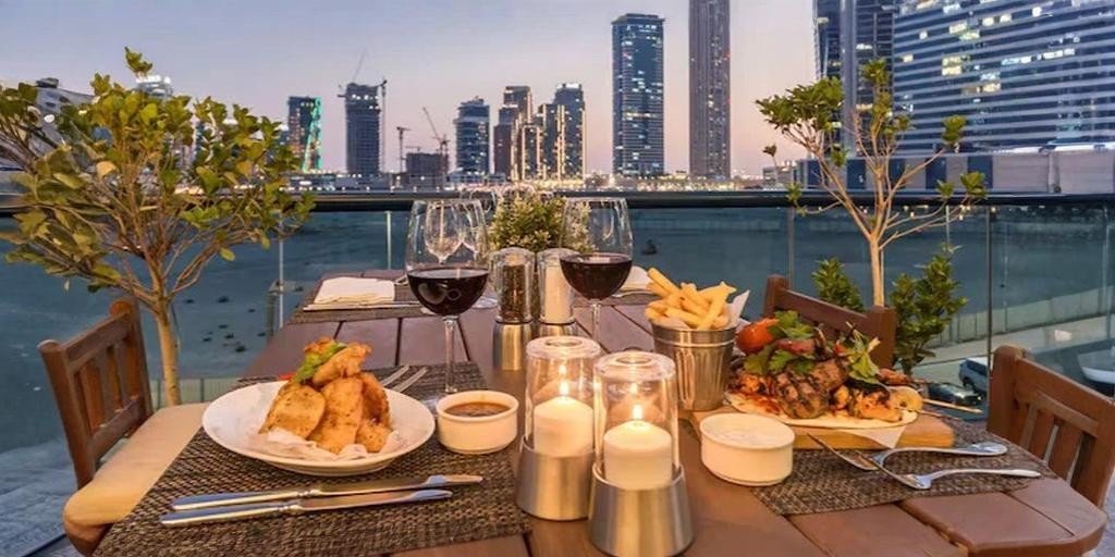 Outdoor beachfront dining table set for two at sunset, featuring plated meals, wine glasses, candles, and a city skyline view across the water.