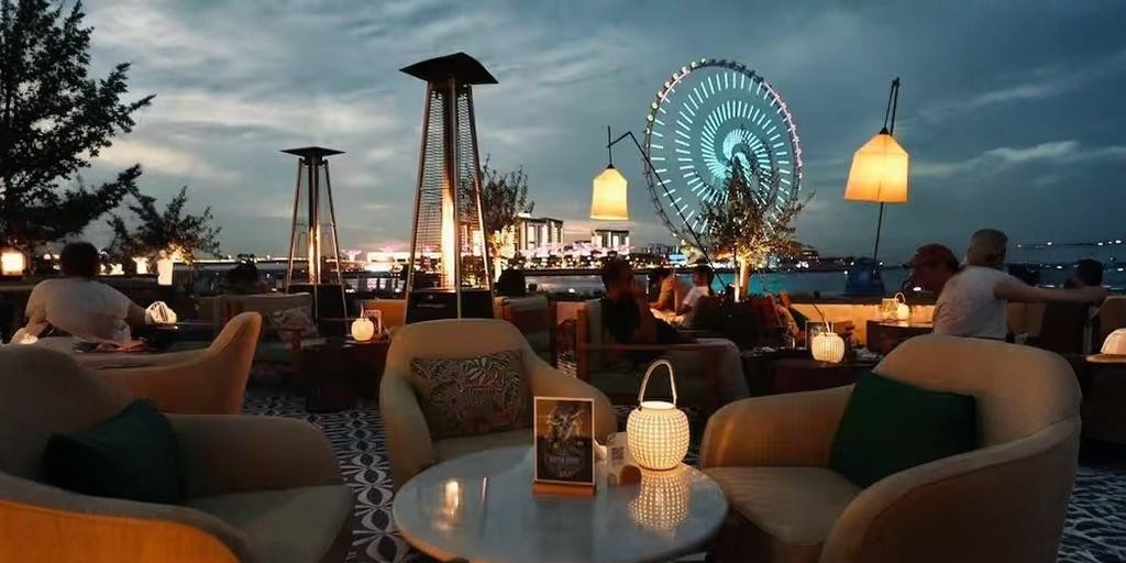 Evening view of an outdoor rooftop lounge with cozy seating, lantern lighting, and guests relaxing, overlooking the waterfront and a brightly lit observation wheel in the distance.