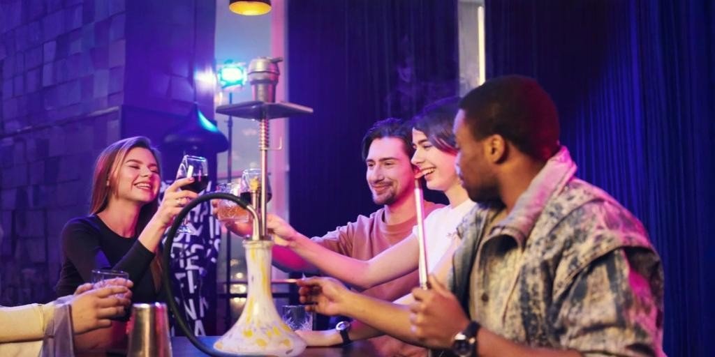 Group of friends enjoying shisha and drinks at a lively indoor lounge, smiling and chatting around a table with colorful lighting and a hookah at the center.