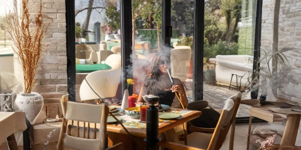 Woman relaxing at an outdoor style café table while smoking shisha, with food and drinks on the table and a garden patio visible through glass doors.