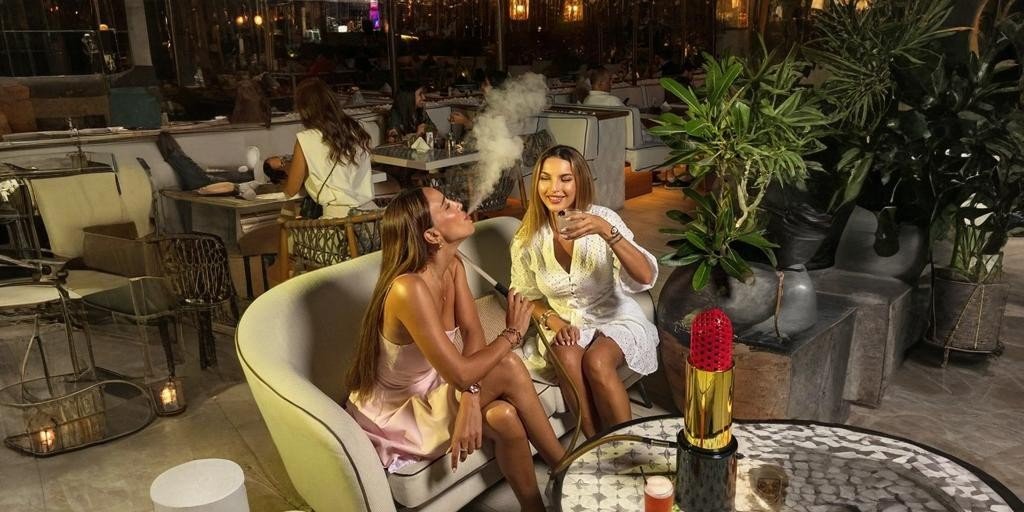 Two women sitting on an outdoor sofa at a stylish shisha lounge in Dubai, enjoying drinks and shisha smoke at night, with warm lighting, plants, and other guests dining in the background.