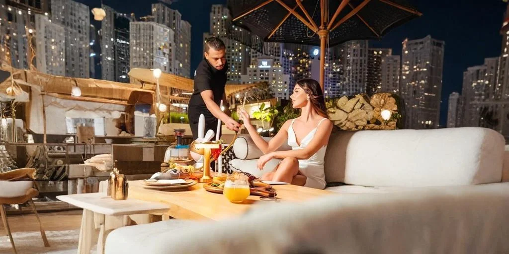 Woman relaxing on an outdoor sofa at a rooftop shisha lounge at night while a waiter serves her, with city skyline lights, umbrellas, and a table set with drinks and shisha in the foreground.